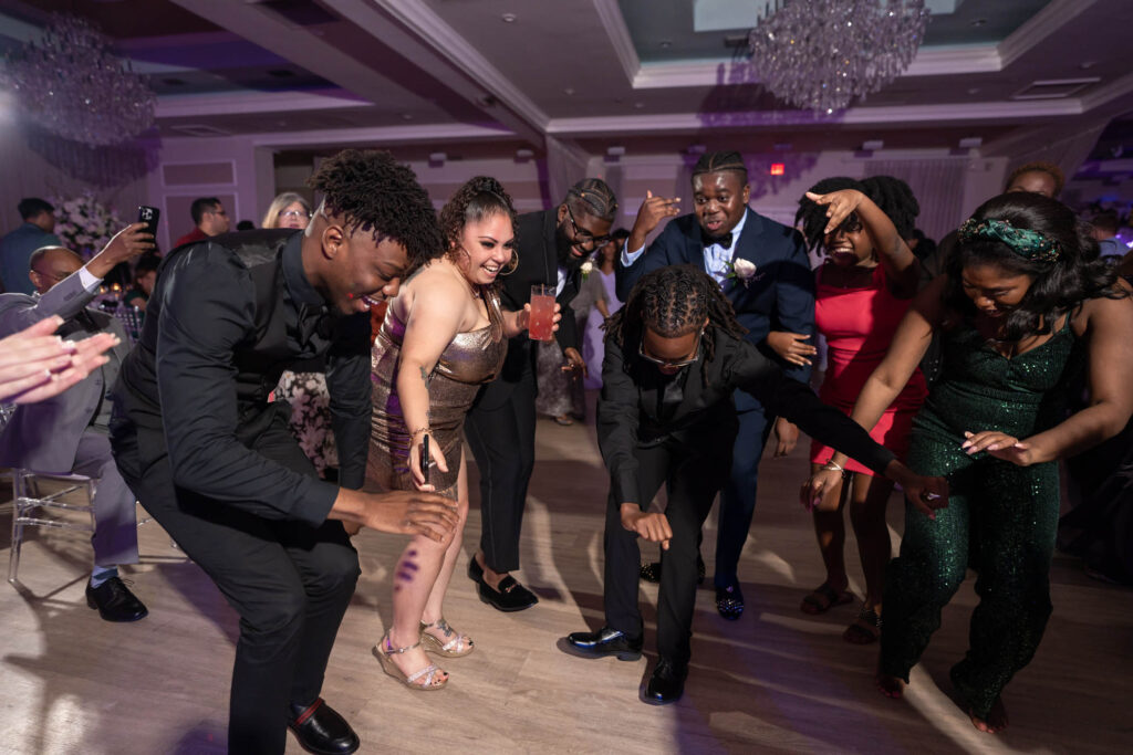 A group of people dressed formally dance energetically together on a ballroom floor under chandeliers at an indoor event.