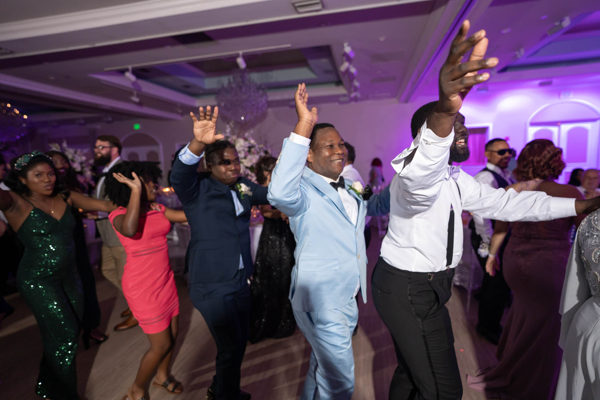 A group of people dressed in formal attire dance in a line at an indoor event with purple lighting.