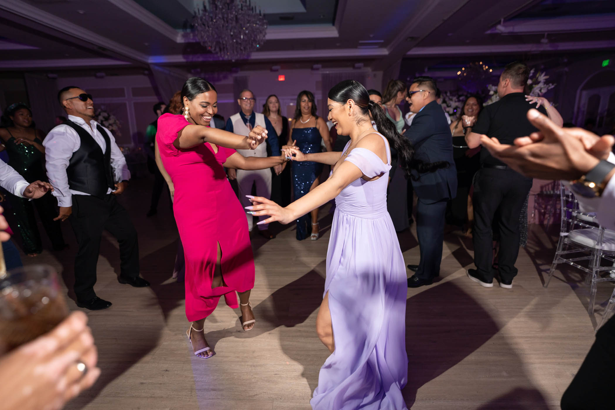 Two women in formal dresses dance together in the center of a lively group at an indoor event, surrounded by other guests on a wooden floor.