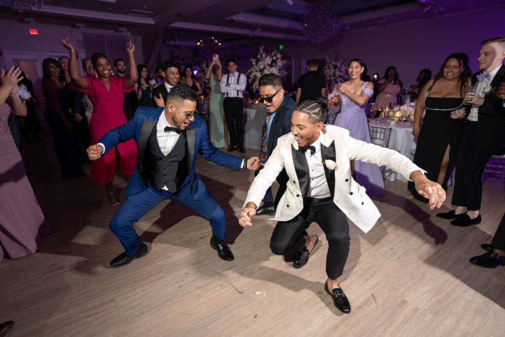 Three men in suits, including one in a white tuxedo jacket, dance energetically on a wooden floor at a formal event while guests watch and cheer in the background.