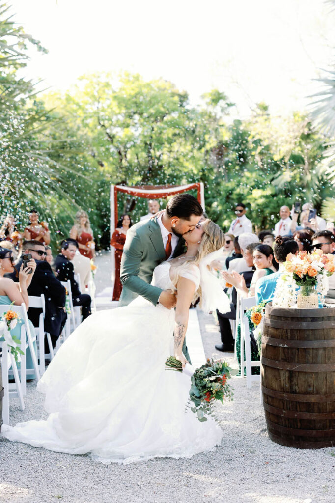 A bride and groom share a kiss at an outdoor wedding ceremony as guests seated on white chairs look on and throw confetti.
