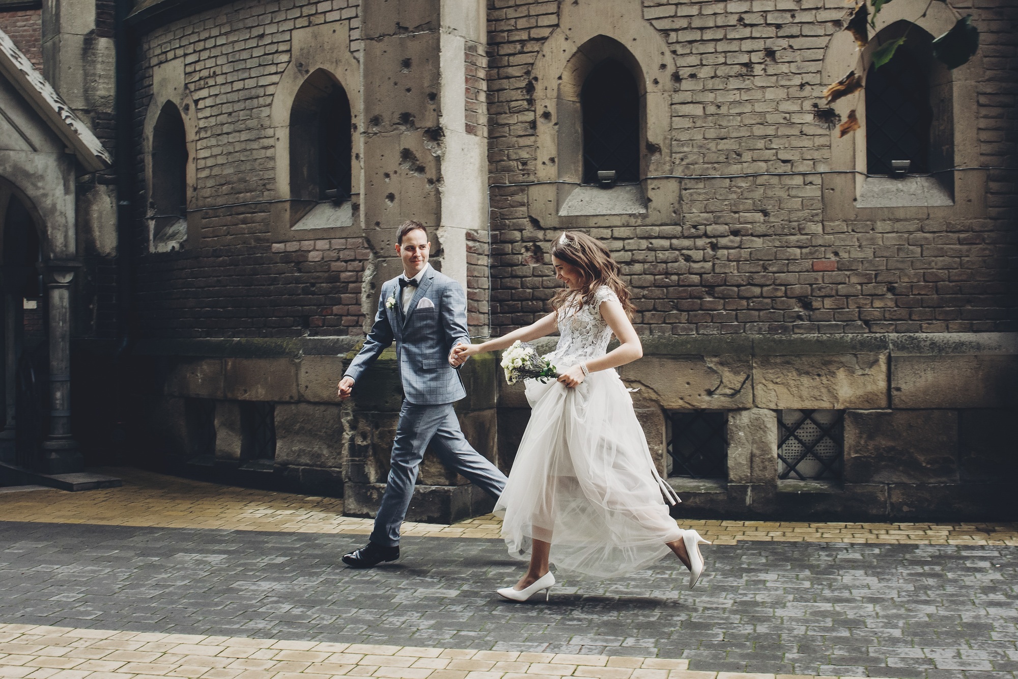 A bride in a white dress and groom in a grey suit walk hand in hand outside a brick building; the bride is holding a bouquet and appears to be mid-step.