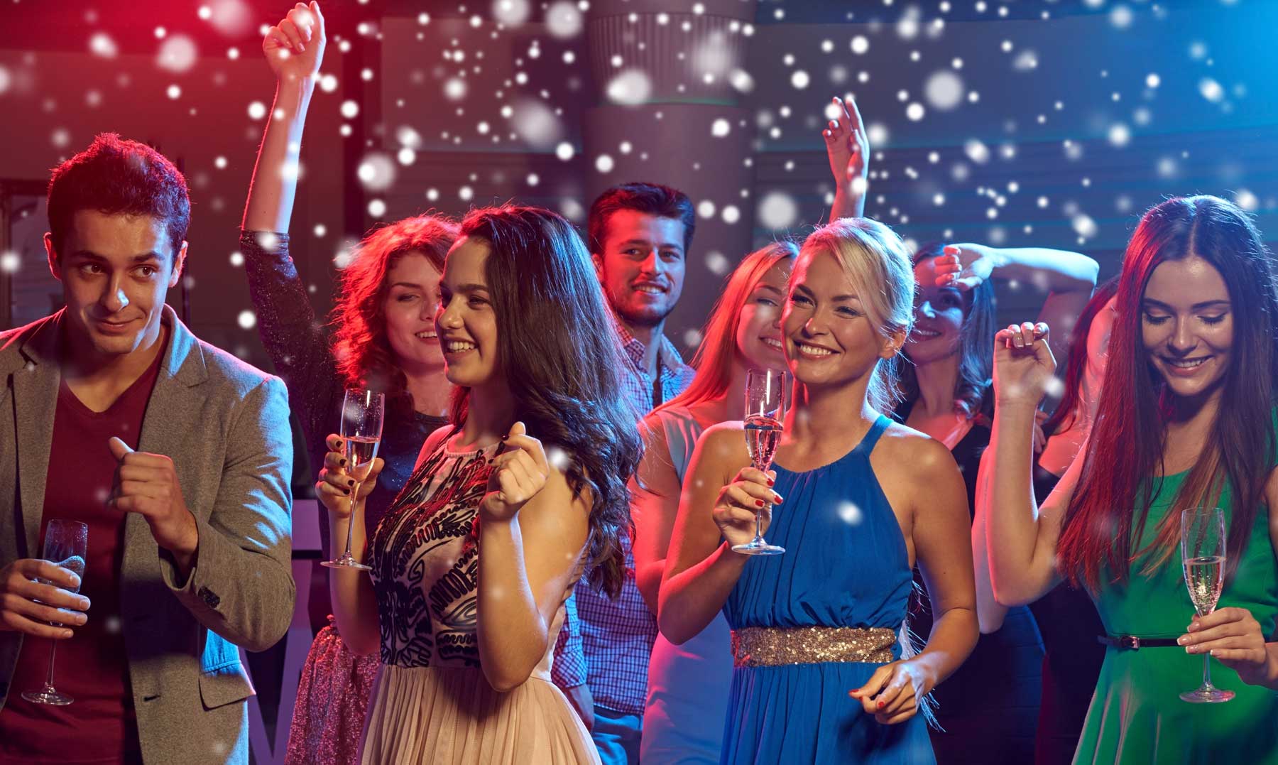 A group of people dressed in party attire dance and hold drinks at a lively indoor celebration with festive lighting.