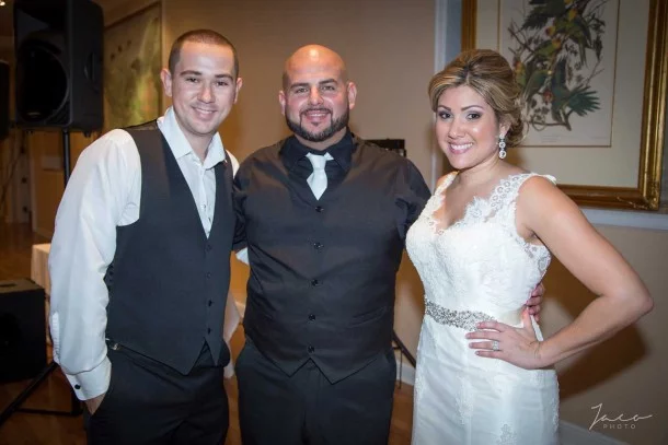 Three people pose together at an indoor event; two men in vests and a woman in a white wedding dress stand smiling for the camera.