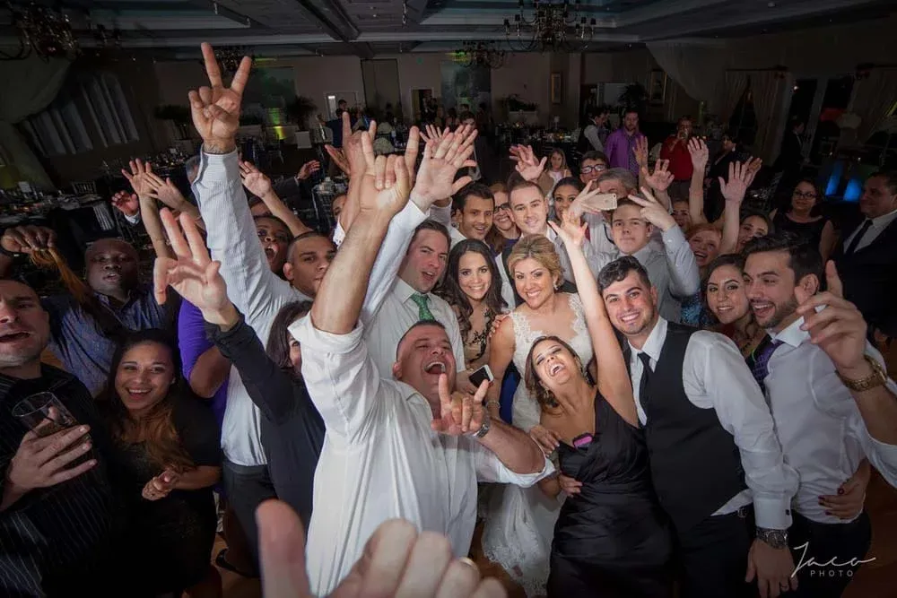 A large group of people dressed formally pose closely together, smiling and raising their hands, likely at a wedding reception.