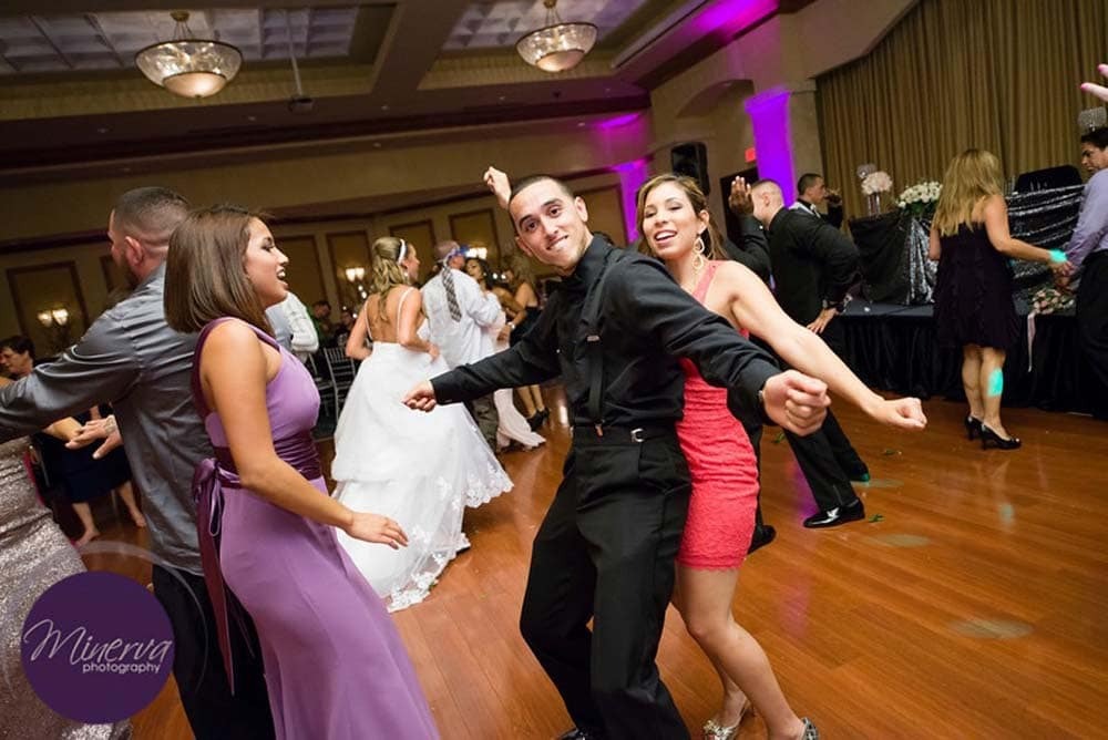People dressed in formal attire dance enthusiastically on a wooden floor at an indoor event, with a bride in the background.