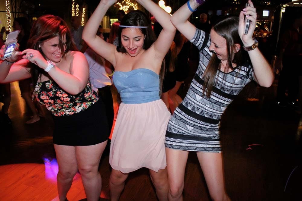 Three young women are dancing together on a dark indoor dance floor, smiling and raising their arms, with festive lights in the background.