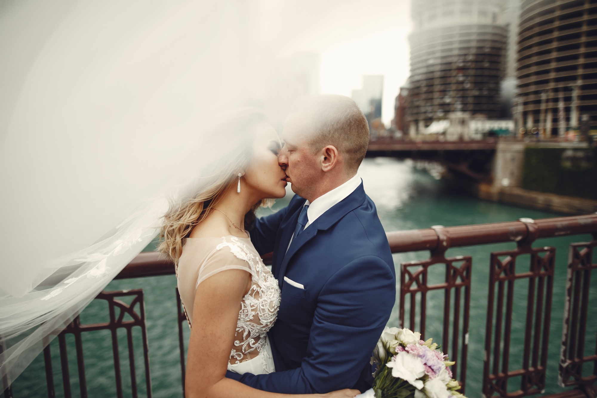 A bride and groom embrace on a bridge by the water, with the bride’s veil flowing in the wind and city buildings visible in the background.