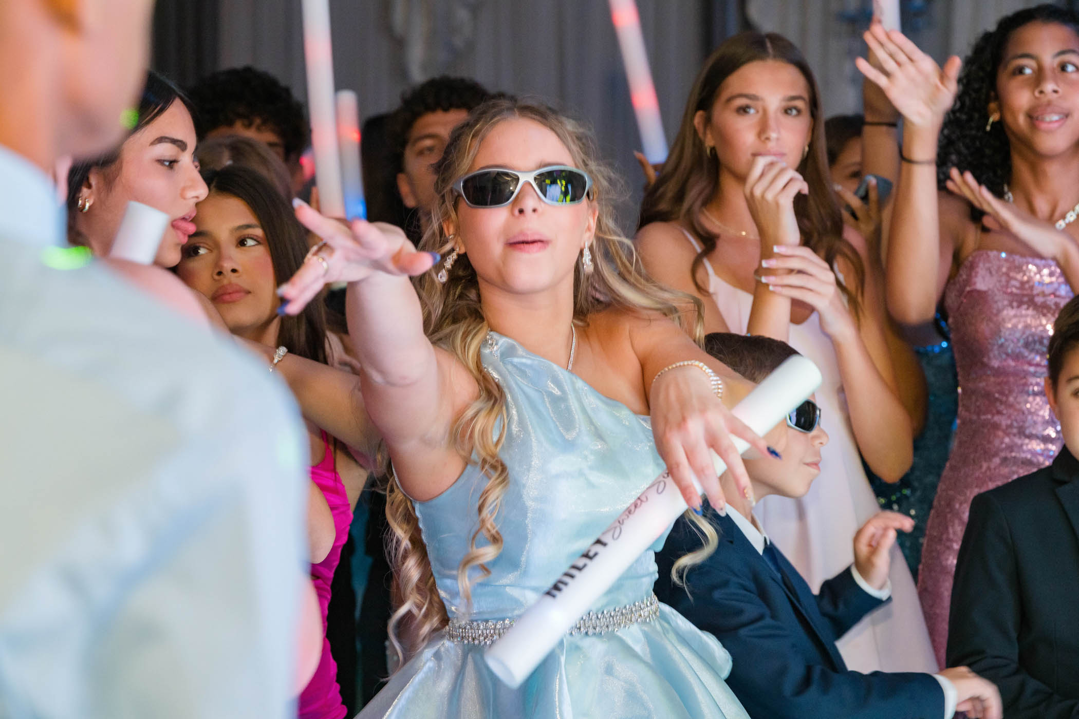 A girl in a light blue dress and sunglasses reaches forward at a crowded party, surrounded by people holding glow sticks.