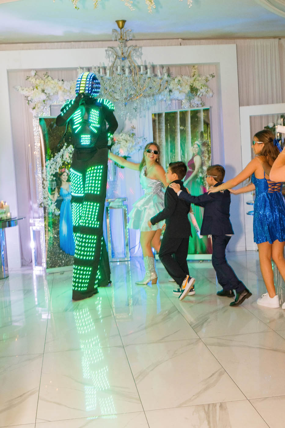 A performer in an LED light suit interacts with children dressed formally on a shiny marble floor at an indoor event.