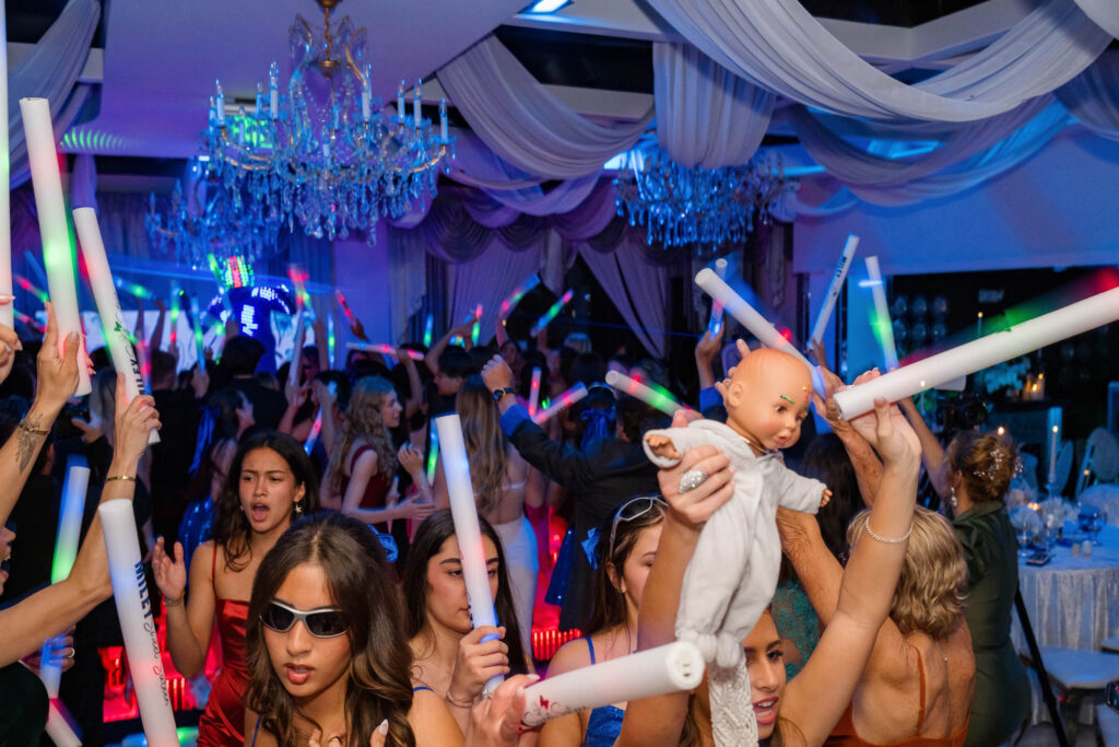 A lively party scene with people dancing and waving glow sticks, while one person in the foreground holds up a baby doll.