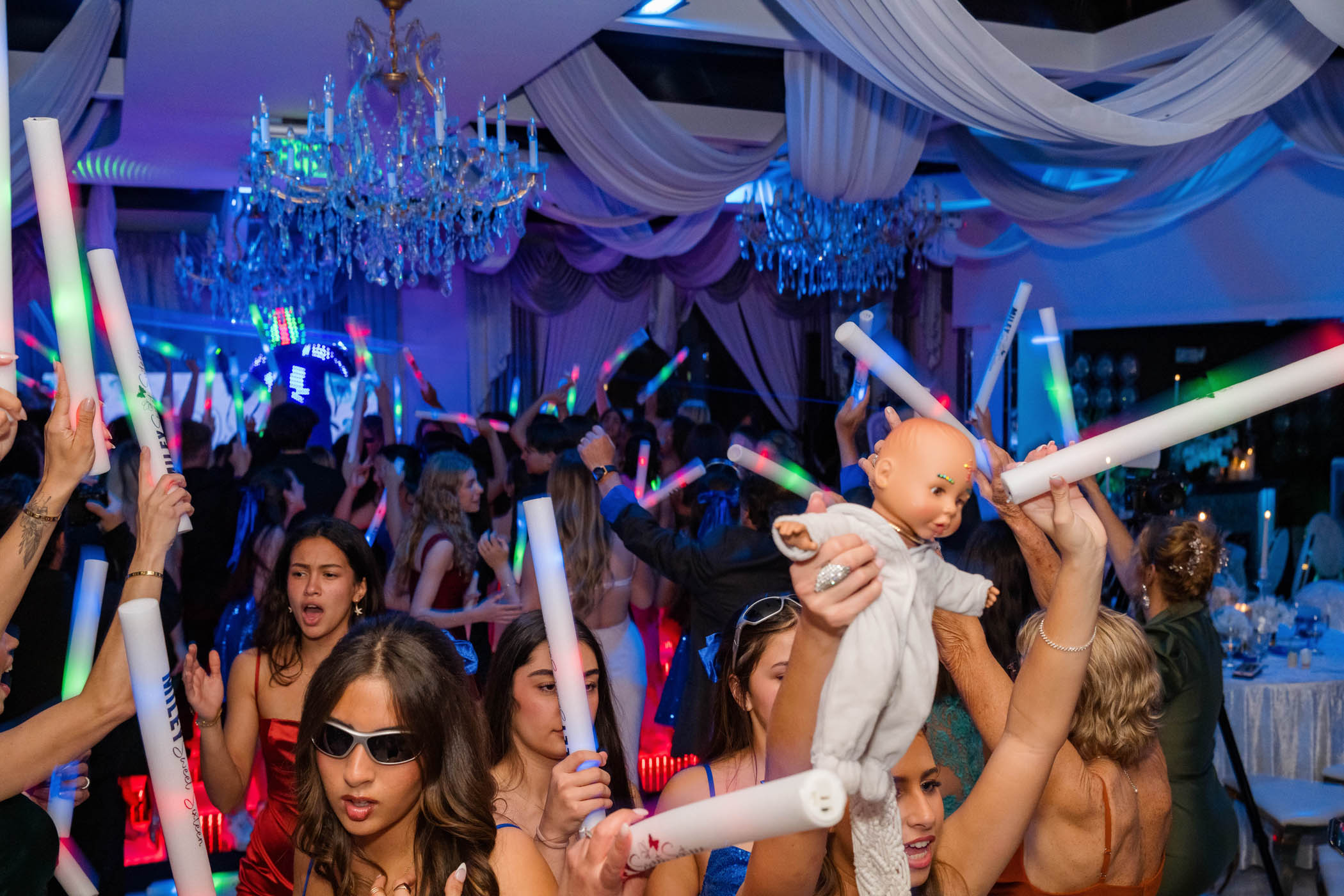 A lively party scene with people dancing and waving glow sticks, while one person in the foreground holds up a baby doll.