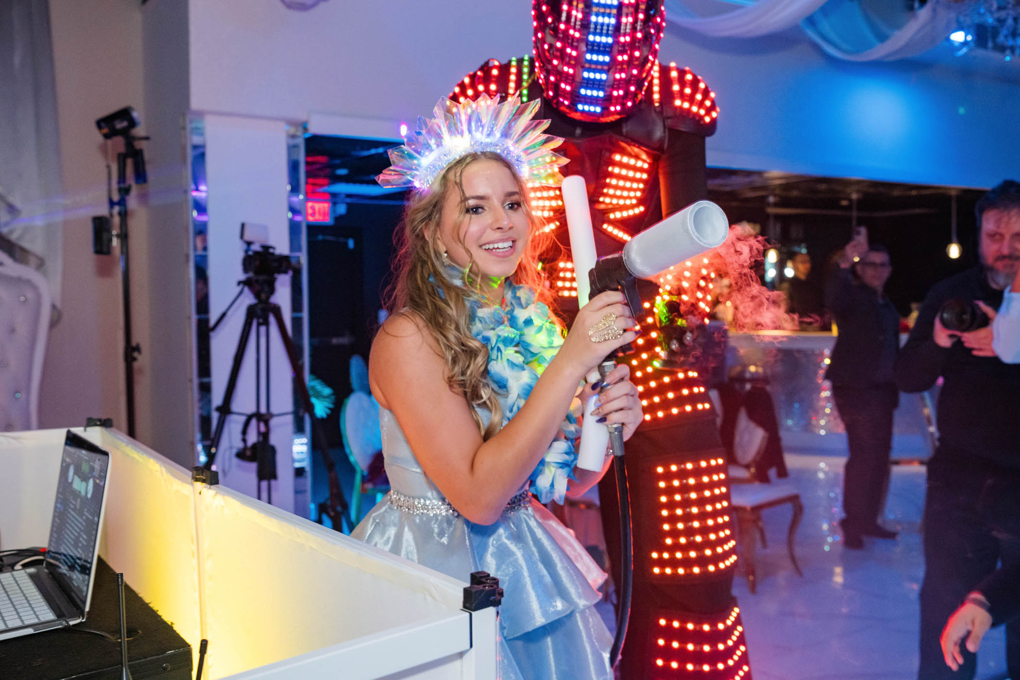 A young woman in a silver dress and light-up crown holds a fog cannon, with a person in an LED robot costume standing behind her at a lively indoor event.
