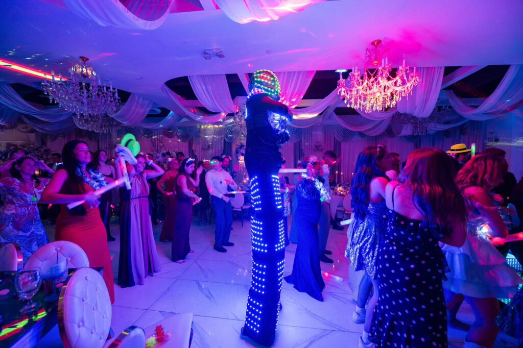 People in formal attire dance and watch a performer in an LED-lit costume on stilts at a party under chandeliers and draped fabric, with colorful lights illuminating the scene.