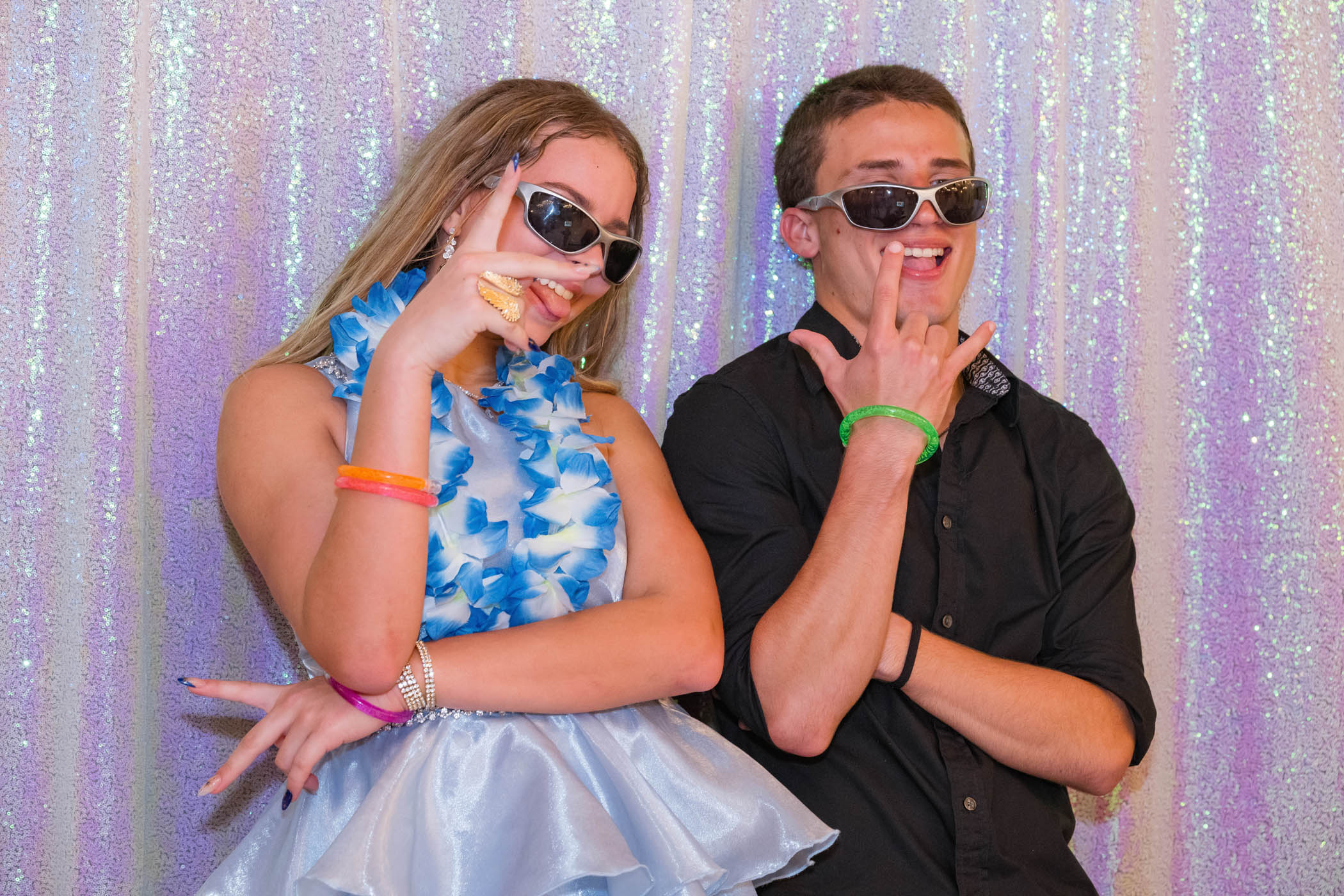 Two young people wearing sunglasses and leis pose playfully in front of a sparkly backdrop, each making hand gestures and smiling.