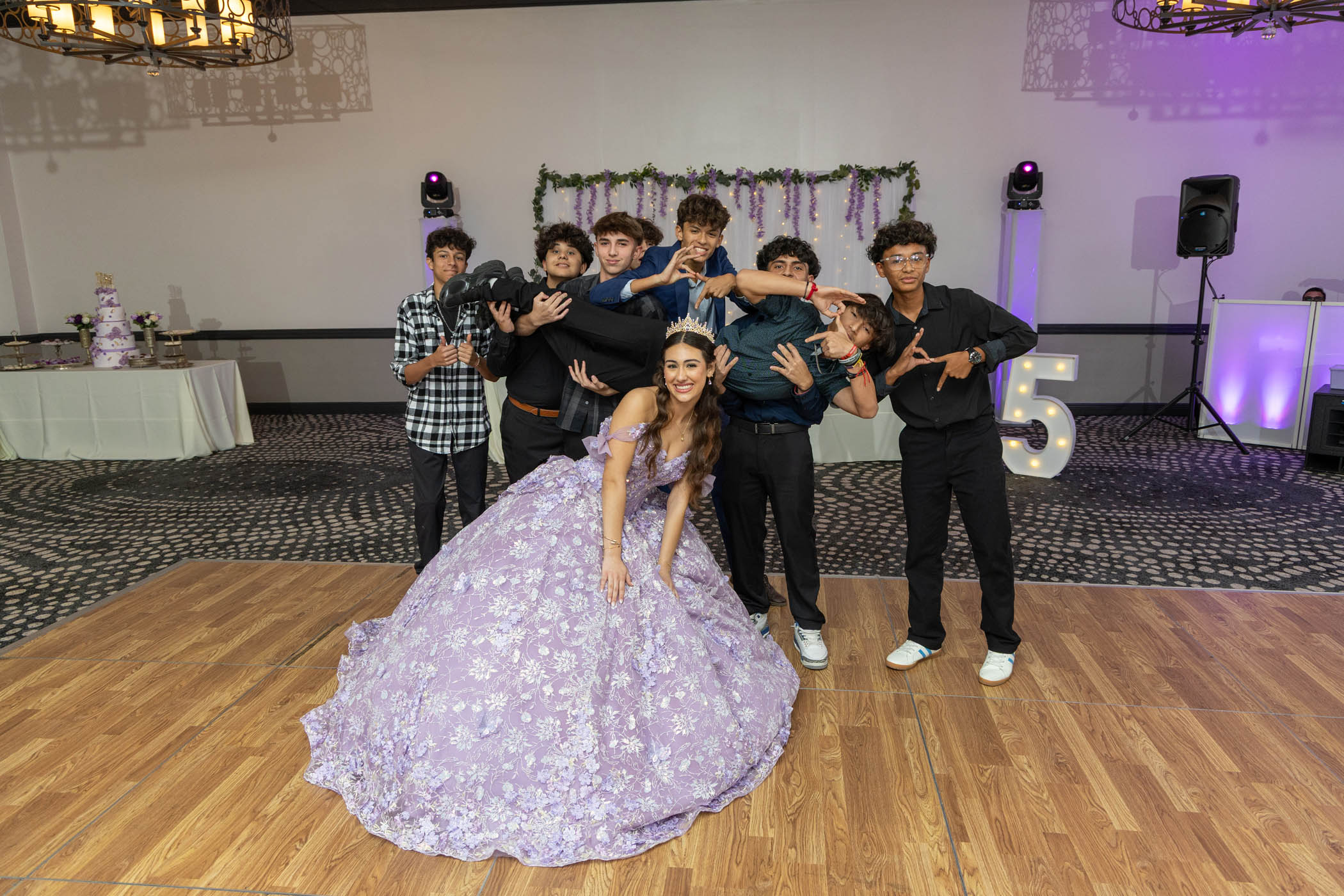 A young woman in a lavender ball gown poses in front of seven young men; one is being held horizontally by the others. Party decorations and a number 15 are in the background.