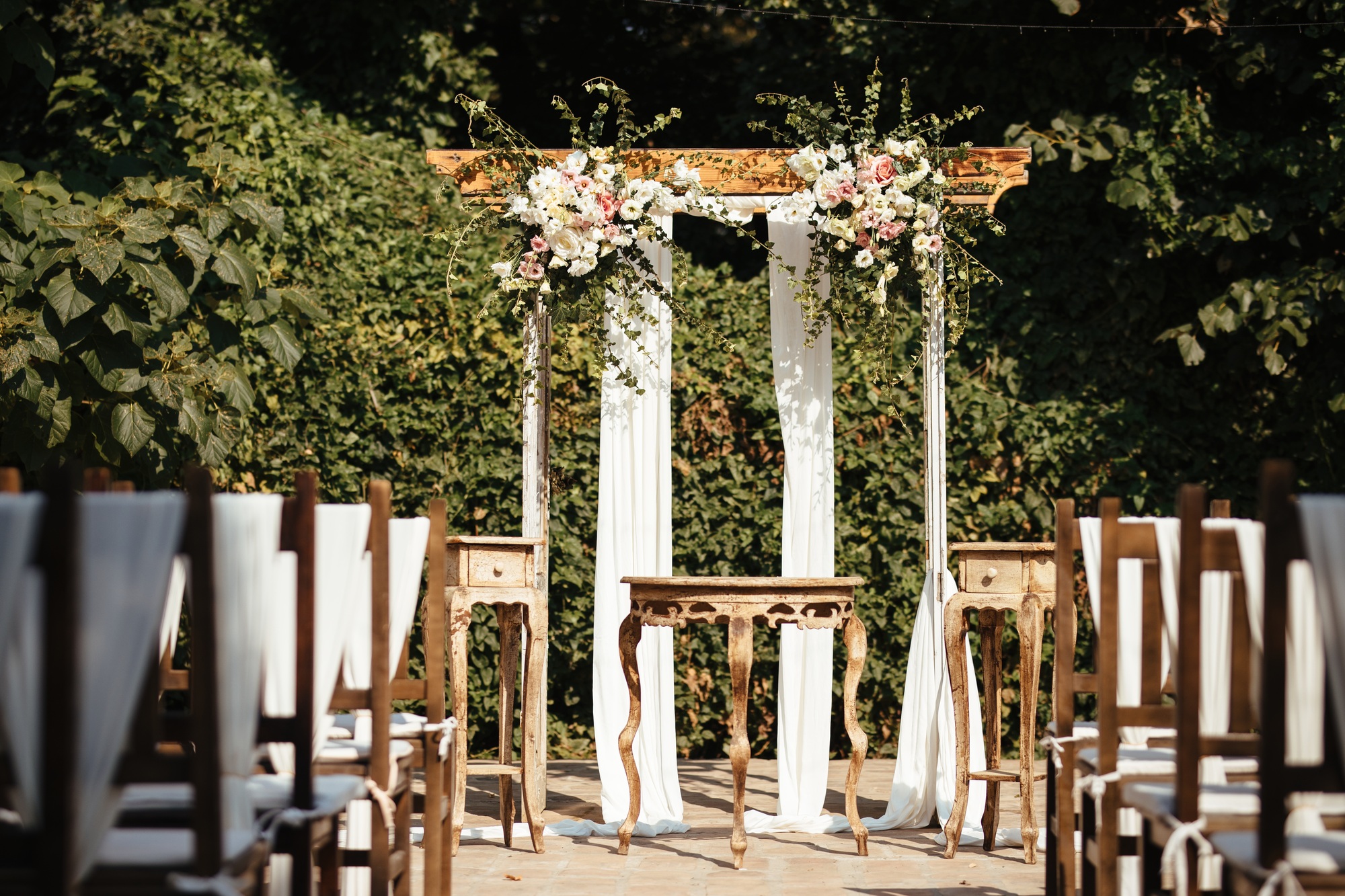 Outdoor wedding ceremony setup with rows of wooden chairs facing a decorated wooden archway adorned with white fabric and floral arrangements, surrounded by greenery.