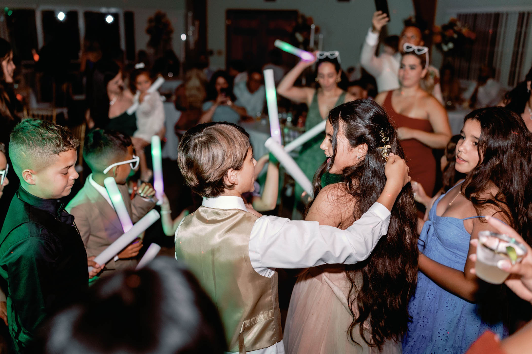 Children and young adults dance together at an indoor event, holding glowing sticks, with people seated and standing in the background.