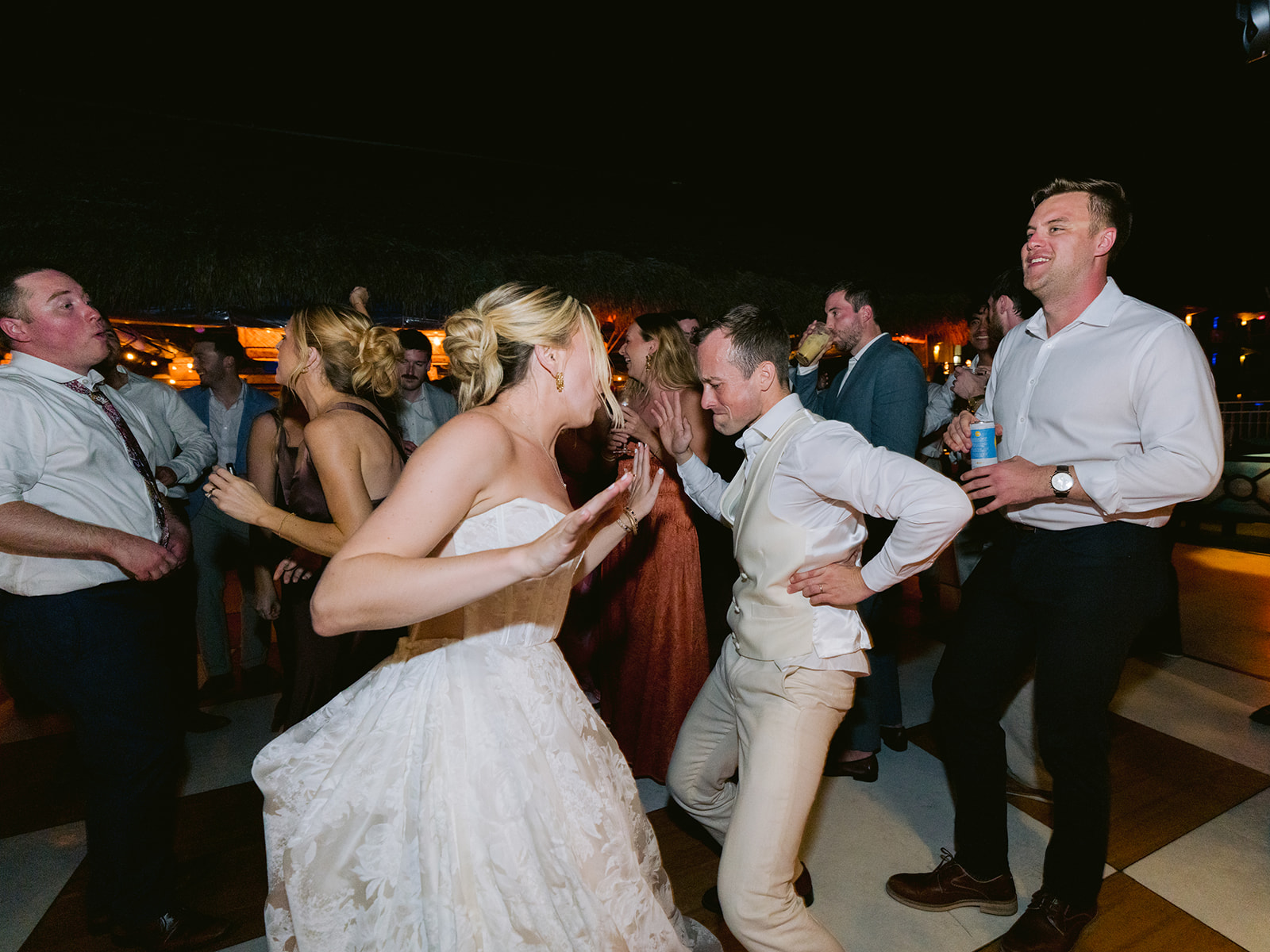 A group of people dance energetically at a party, with a bride in a white dress and a man in a vest and tie in the foreground.