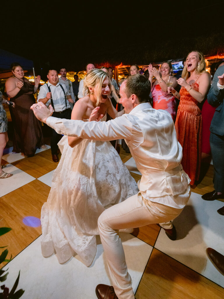 A bride and groom dance energetically together on a checkered floor, surrounded by cheering guests at a wedding reception.