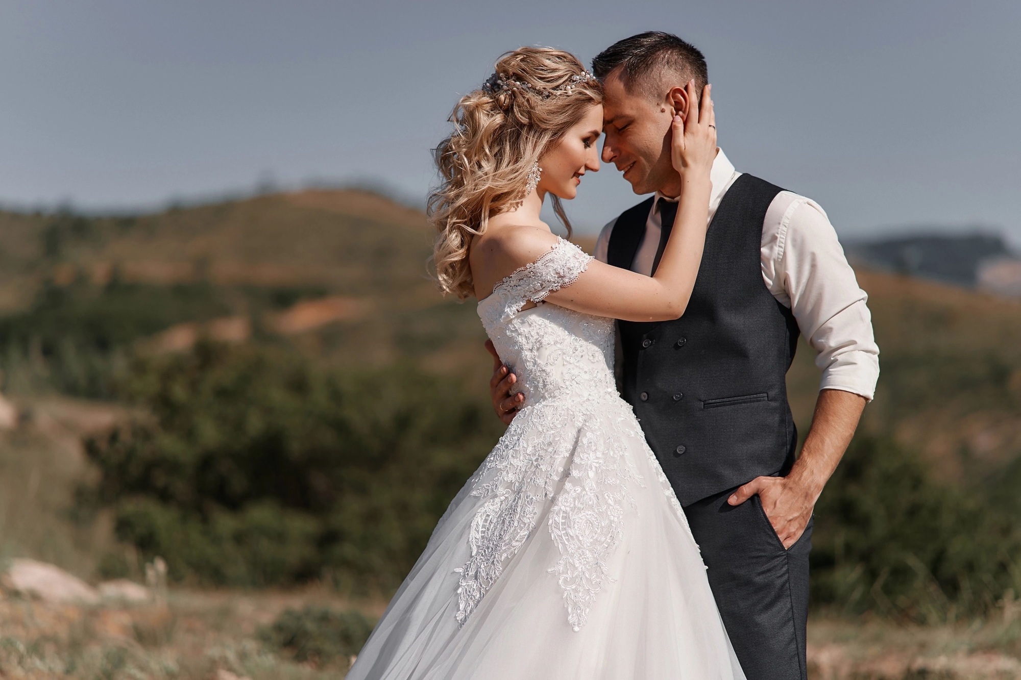 A bride in a white wedding dress and a groom in a vest and tie embrace outdoors with green hills in the background.