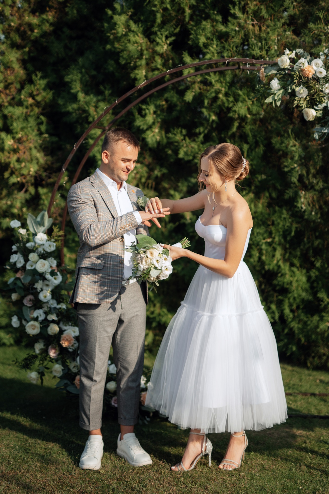A bride and groom stand outdoors exchanging rings under a floral arch, surrounded by greenery and flowers.