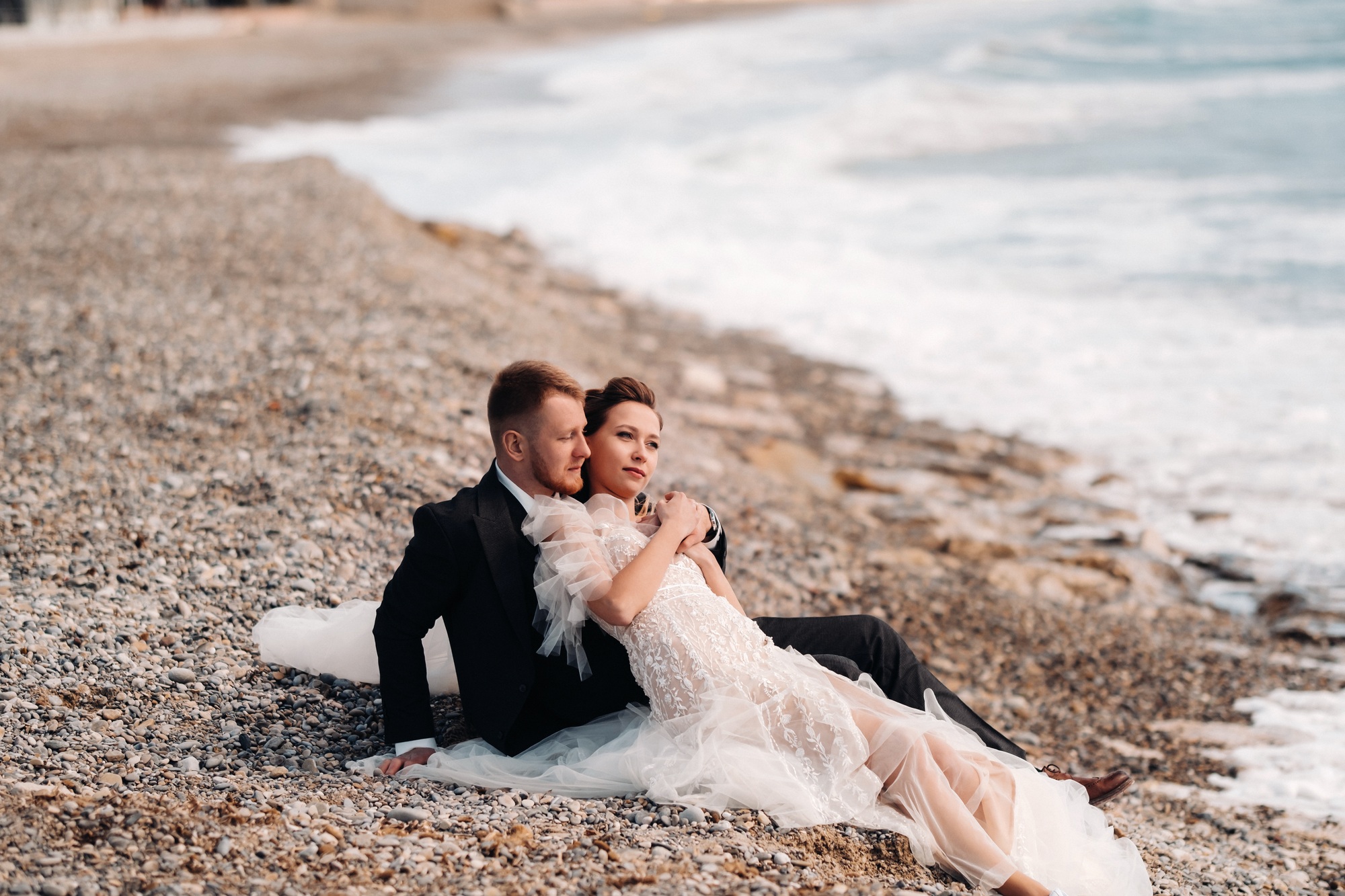 A bride and groom in formal wedding attire sit closely together on a rocky beach near the shoreline, with waves in the background.