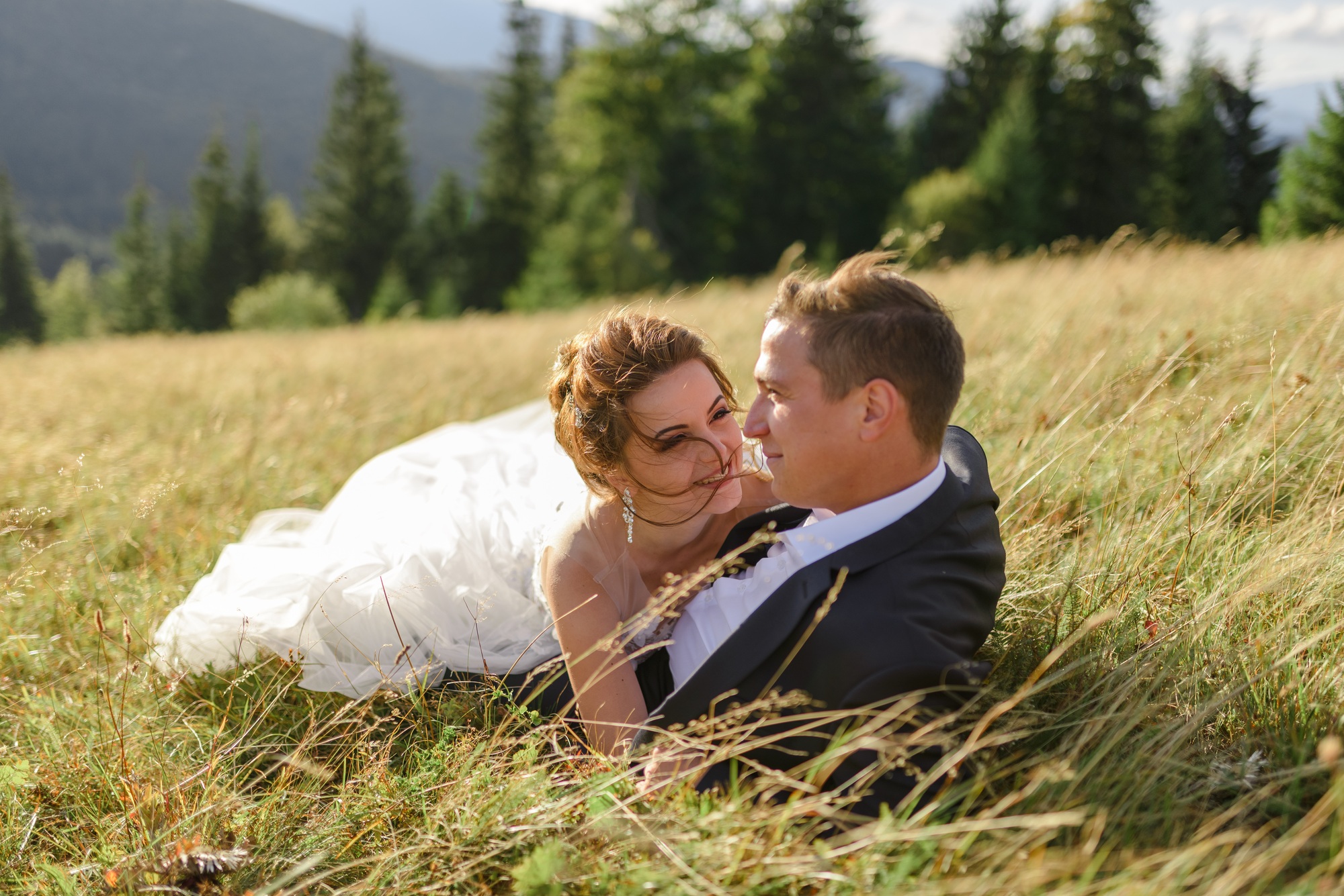 A bride and groom lie in tall grass in a sunlit field, facing each other and smiling, with trees and mountains in the background.