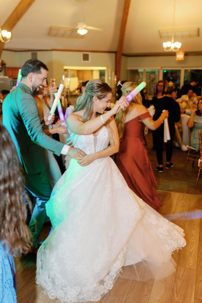 A bride in a white gown dances with a groom in a green suit at a reception, both holding glowing sticks as guests watch and join in the celebration.