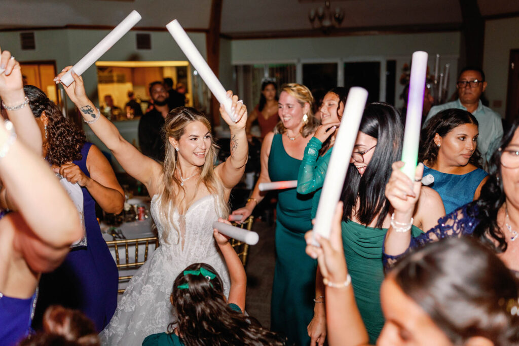 A bride in a white dress celebrates with guests, all holding glowing foam sticks and dancing at a lively indoor wedding reception.
