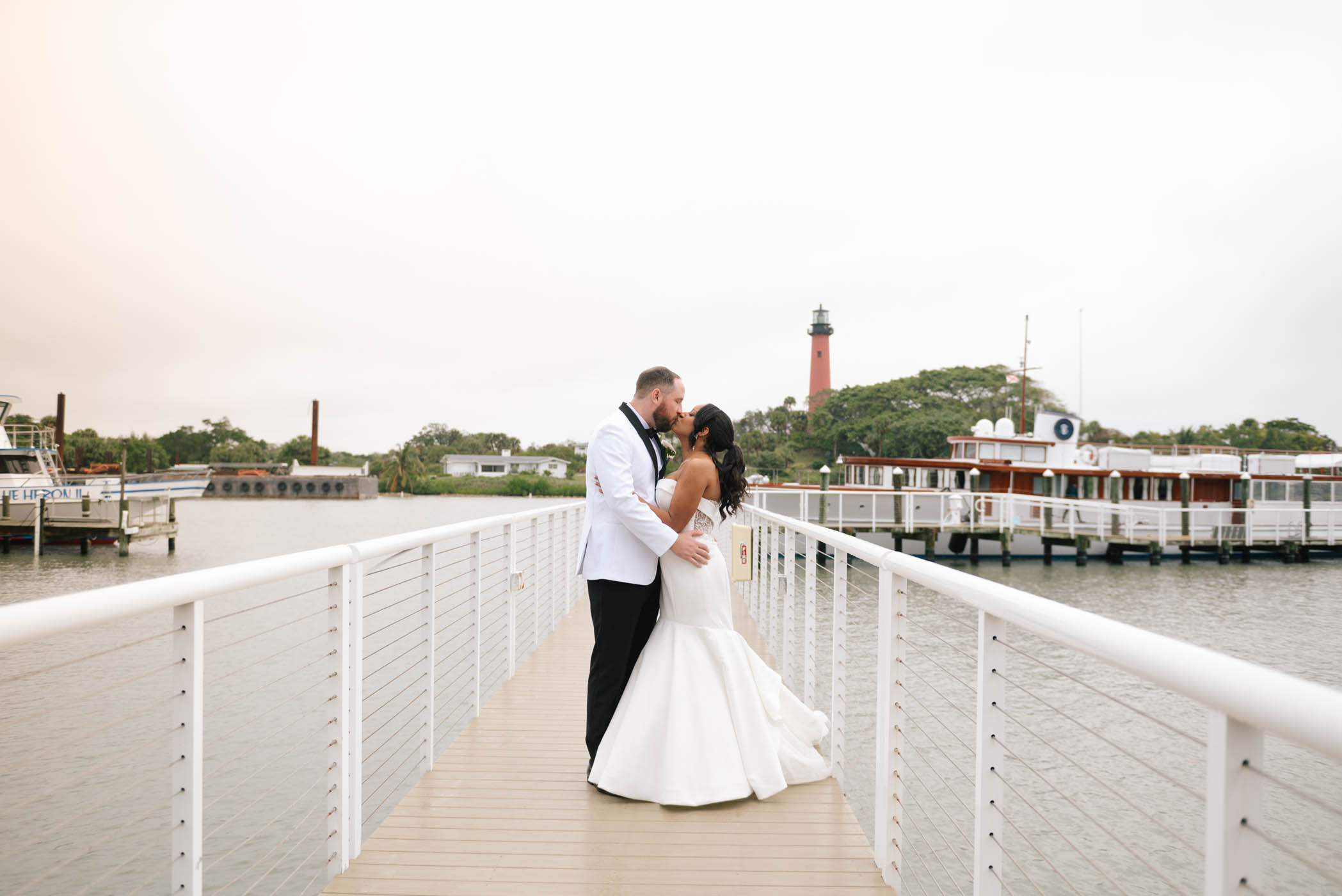 A bride and groom stand on a dock, embracing and looking at each other, with a lighthouse and boats in the background.