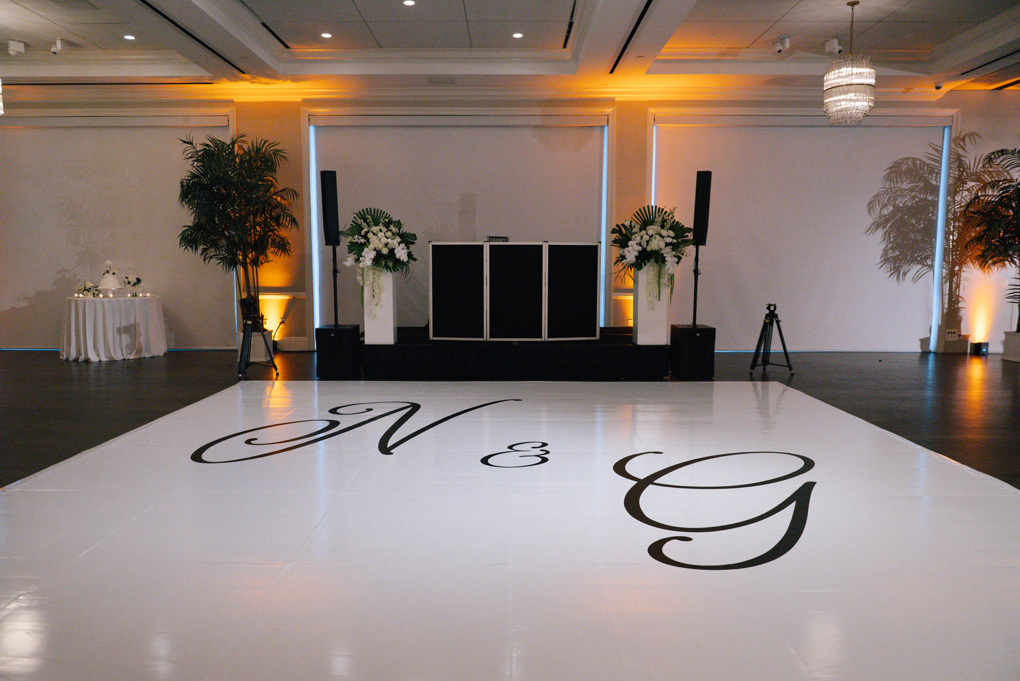 A decorated event space with a white dance floor featuring large black letters "N & G," a DJ booth, speakers, and floral arrangements.