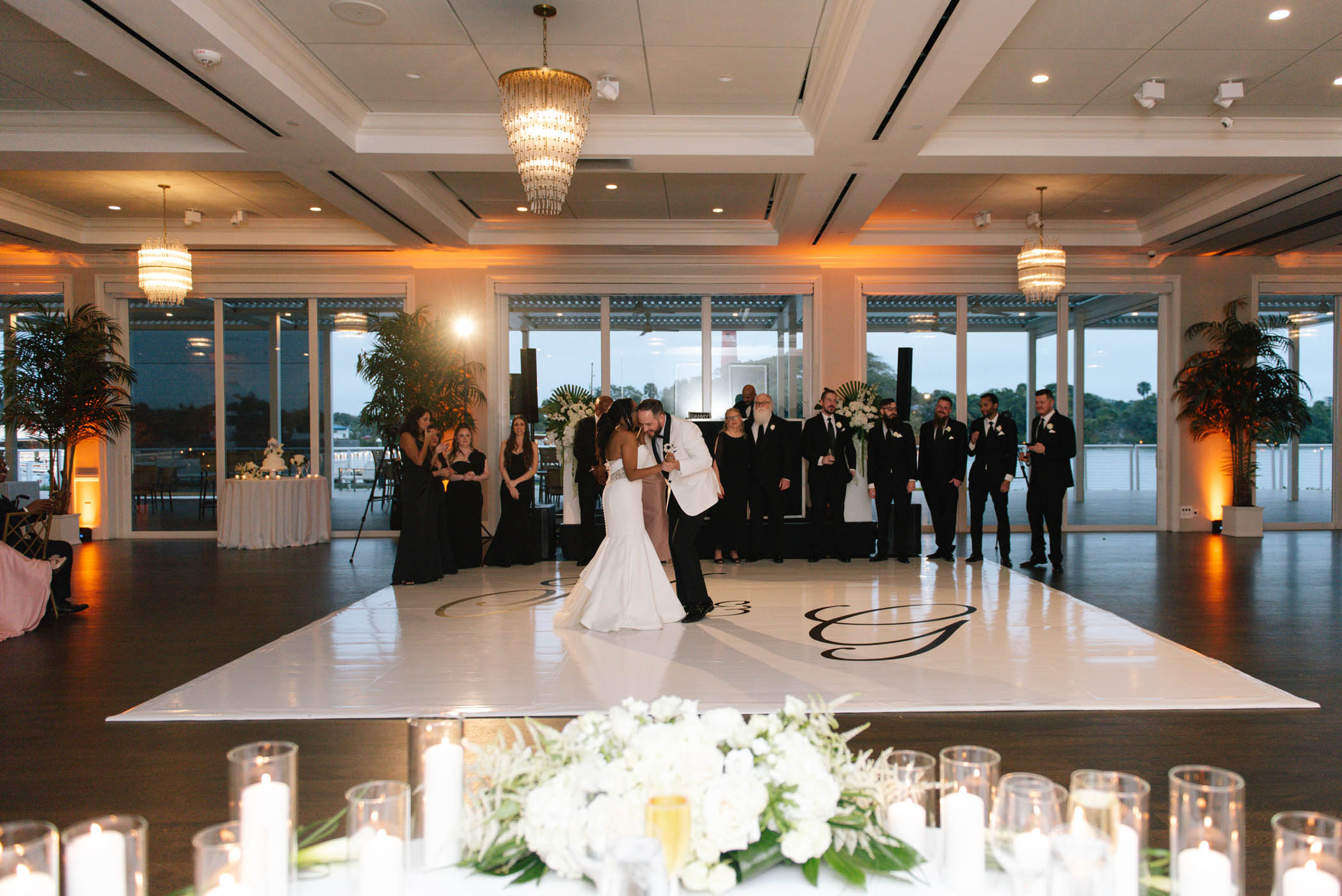 A bride and groom dance on a white floor in a ballroom, surrounded by wedding guests dressed in formal attire. Large windows and floral arrangements are visible in the background.