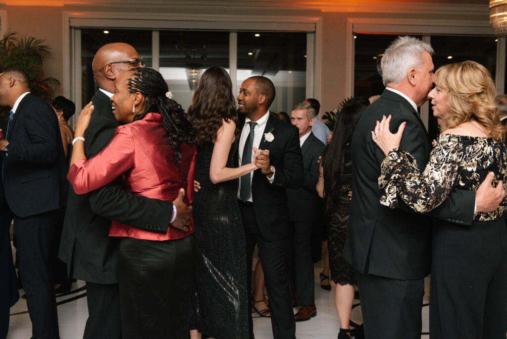 People in formal attire are dancing in pairs at an indoor event, with some smiling and mingling in the background.