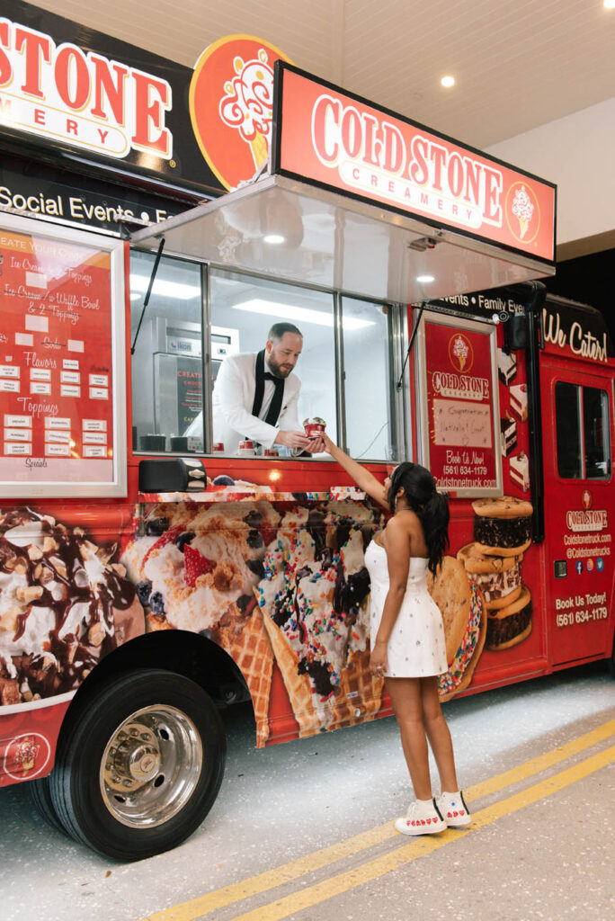 A woman in a white dress receives ice cream from a man inside a Cold Stone Creamery food truck decorated with images of ice cream and treats.