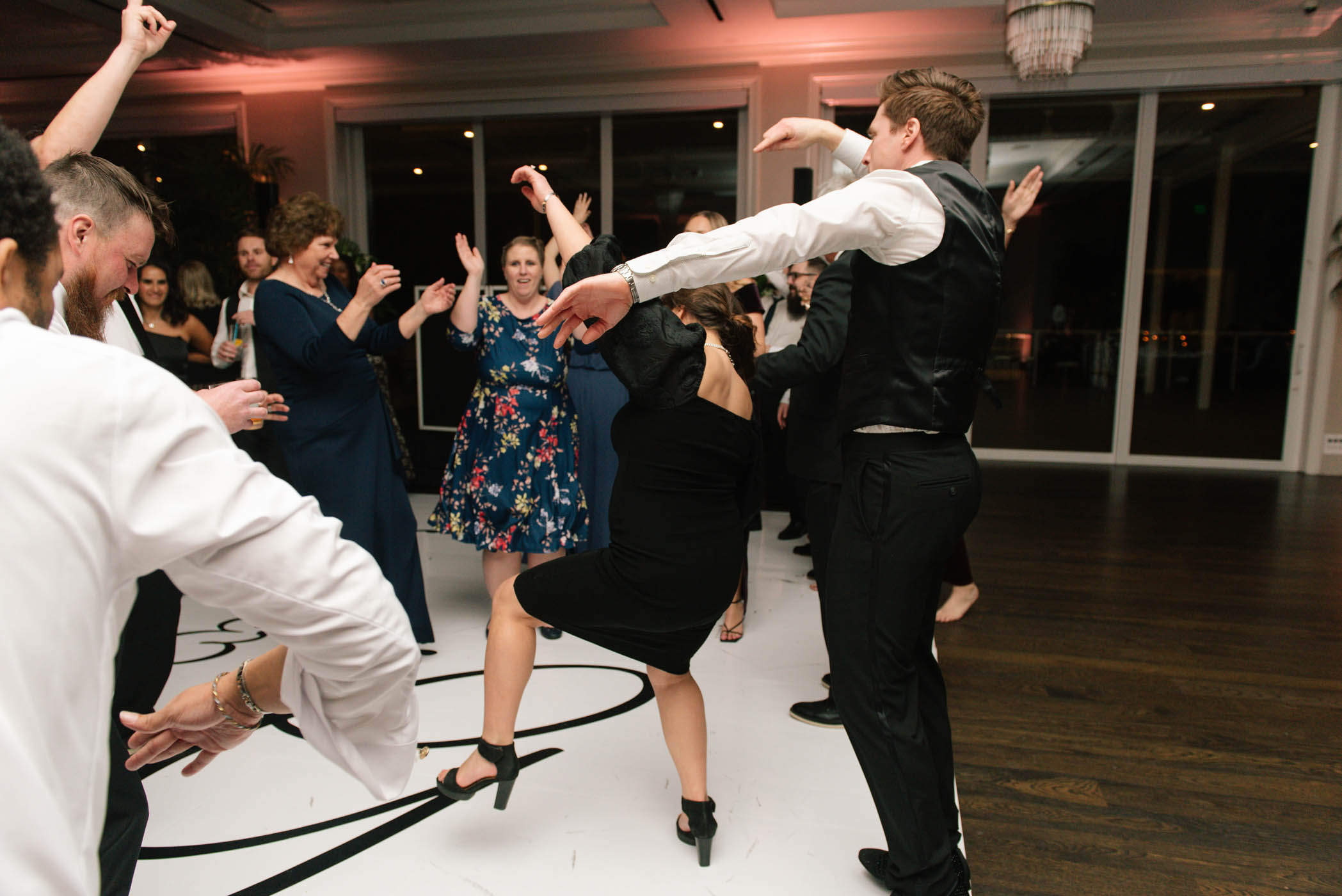 A group of people dressed in formal attire dance energetically on a white floor at an indoor event or party.